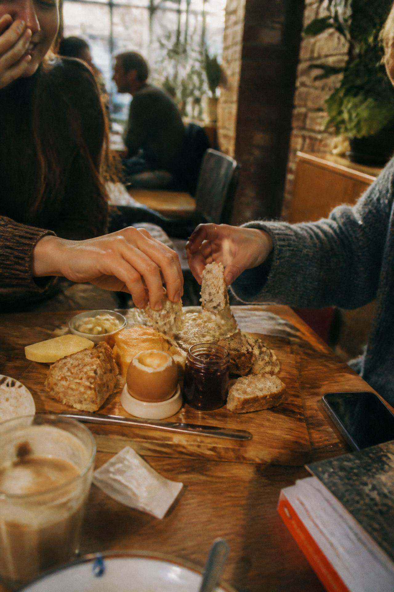 Zwei Personen teilen sich ein Frühstücksbrett mit Brot, Ei, Käse und Marmelade am Cafétisch