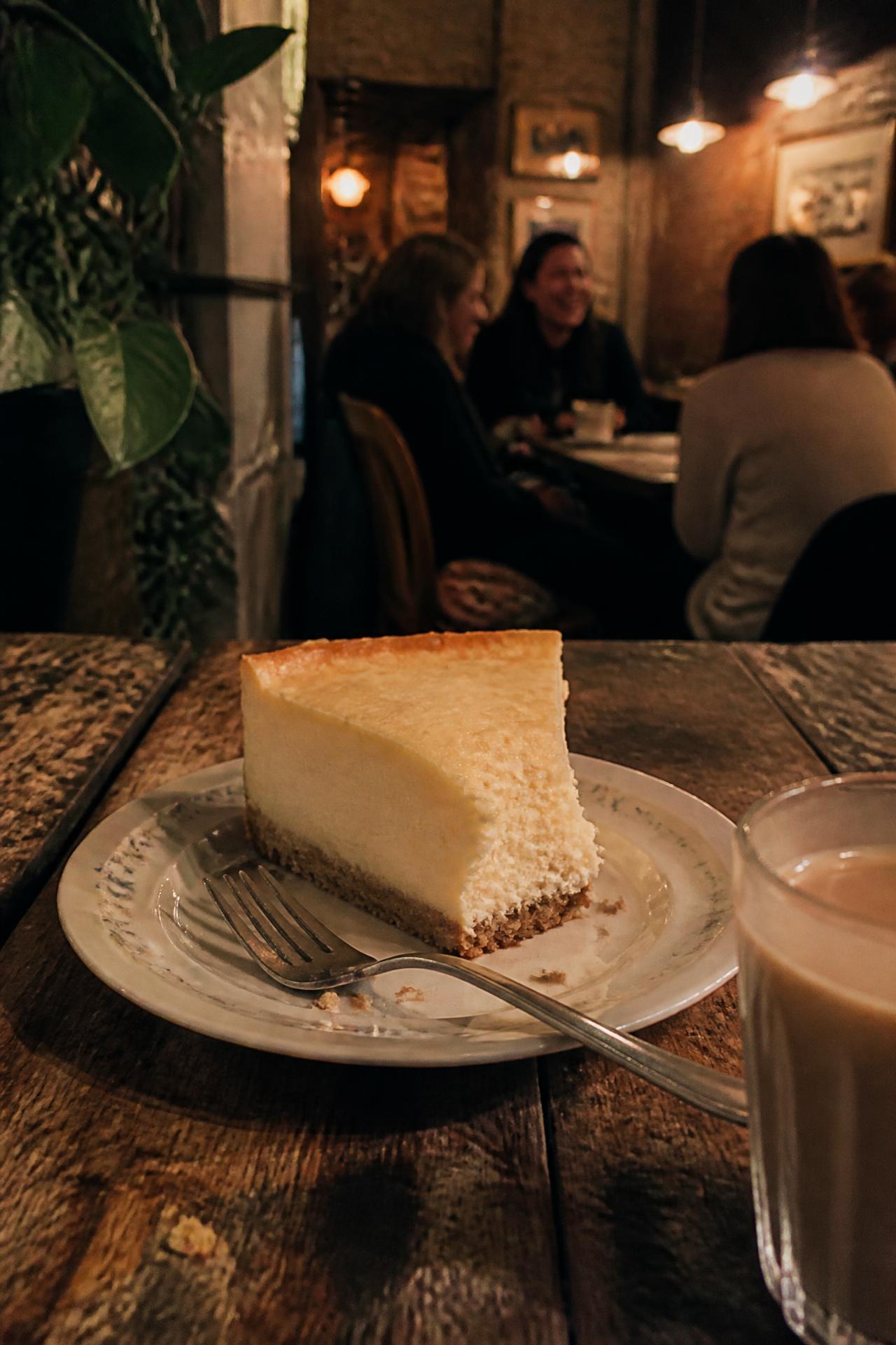 Stück Käsekuchen mit Gabel auf altem Holztisch, im Hintergrund lachende Gäste im warmen Licht