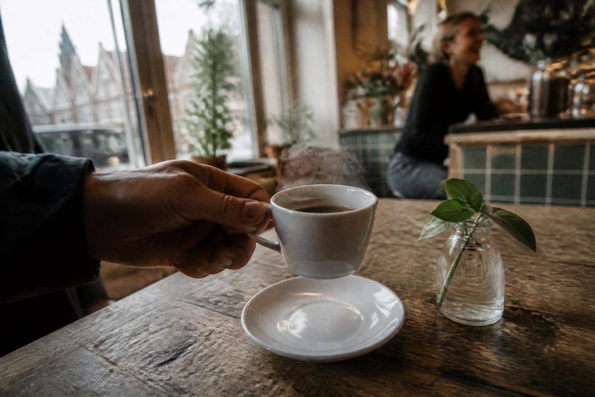 Hand hebt eine Tasse Filterkaffee vom Holztisch, Dampf im Fensterlicht