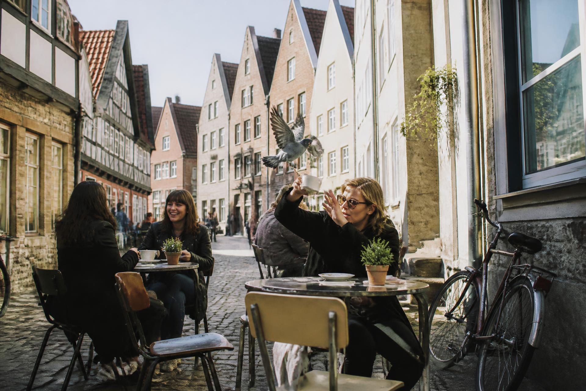 Tische auf dem Kopfsteinpflaster der Altstadtgasse, eine Gästin verscheucht eine Taube von der Tasse