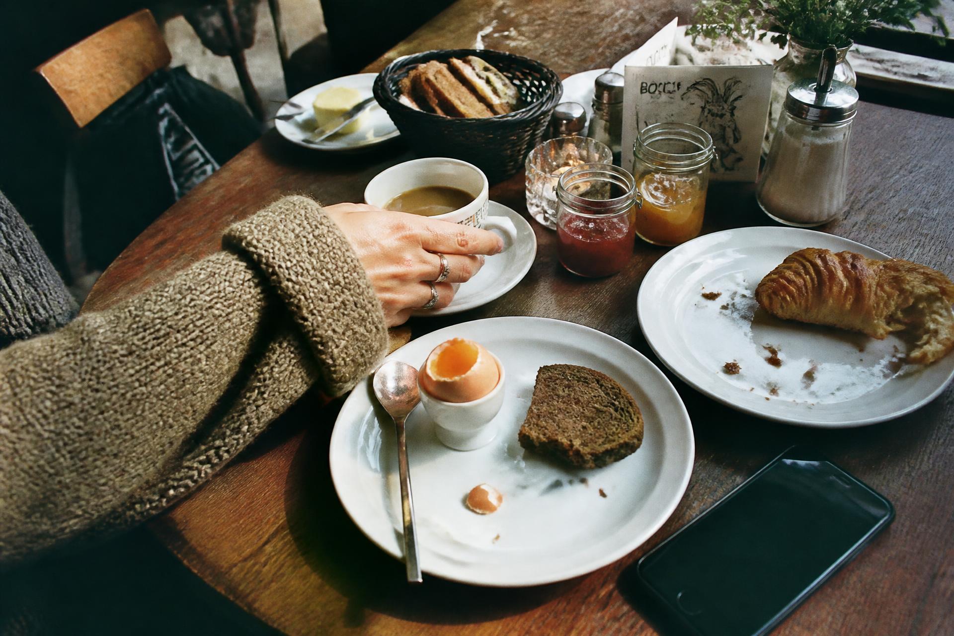 Gedeckter Frühstückstisch mit weichem Ei, dunklem Brot und Marmeladengläsern, eine Hand im Strickpullover greift zur Kaffeetasse