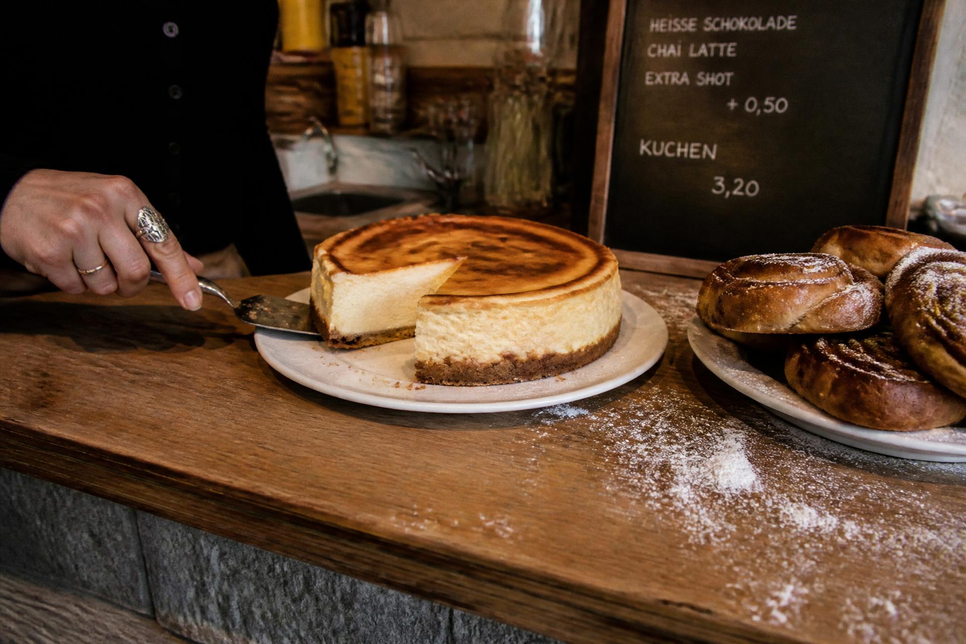 Käsekuchen mit herausgeschnittenem Stück auf dem Tresen, daneben Zimtschnecken und Puderzucker